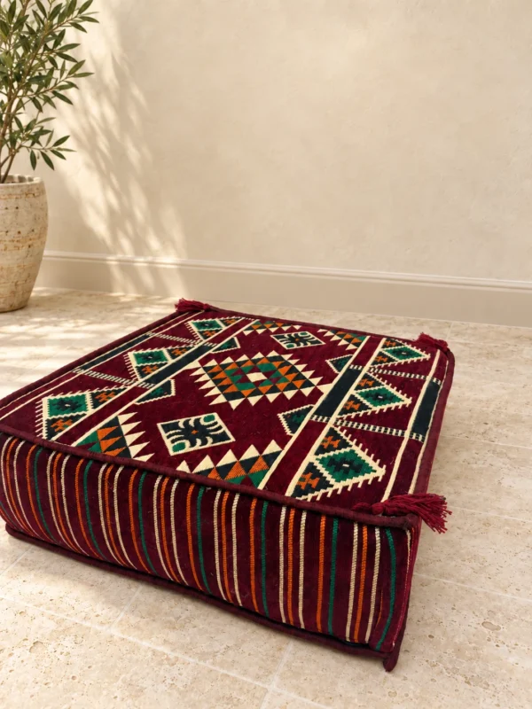 Burgundy kilim-pattern floor pouf with cream, green and orange geometric motifs, striped sides and corner tassels, on a stone floor.