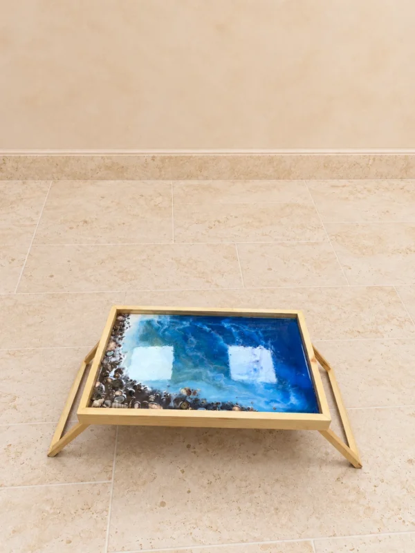 Folding wooden tray table with a blue ocean resin art top, white foam swirls and a shoreline of small shells and pebbles, on a stone floor.