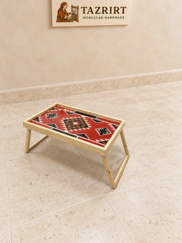 Folding wooden tray table with a red kilim-pattern top, black and cream diamond motifs, on a stone floor.