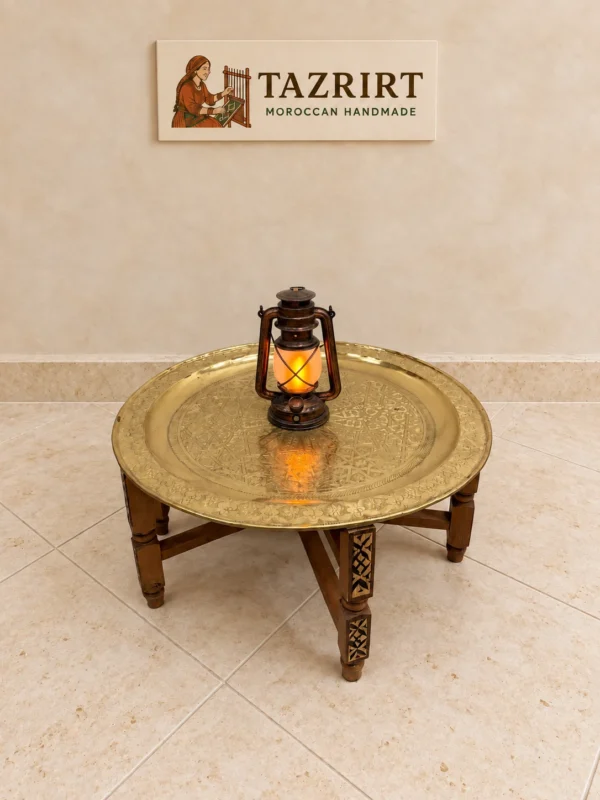 Round hand-engraved Moroccan brass tray table on a folding carved wooden stand, with a small lantern on top, on a stone floor.