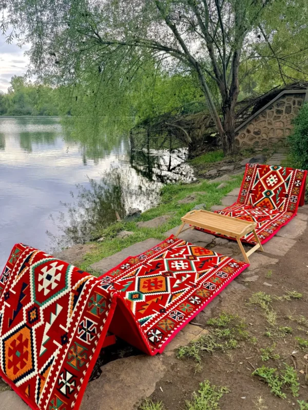 Two red Sahrawi folding outing seats with a kilim-style pattern set by a lake, backrests raised, with a small folding tray between them