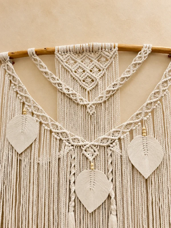 Front detail of the ivory macramé wall hanging: diamond knot panel, braided swags, leaf pendants with wooden beads and tassels.