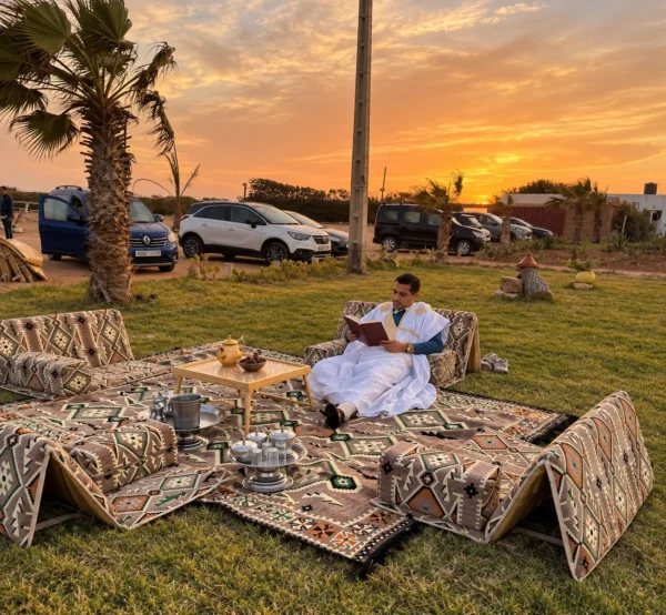 Taupe Sahrawi outing majlis set laid out on grass at sunset: four folding seats with armrest bolsters around a kilim-pattern rug, a man in a white daraa reading on one seat