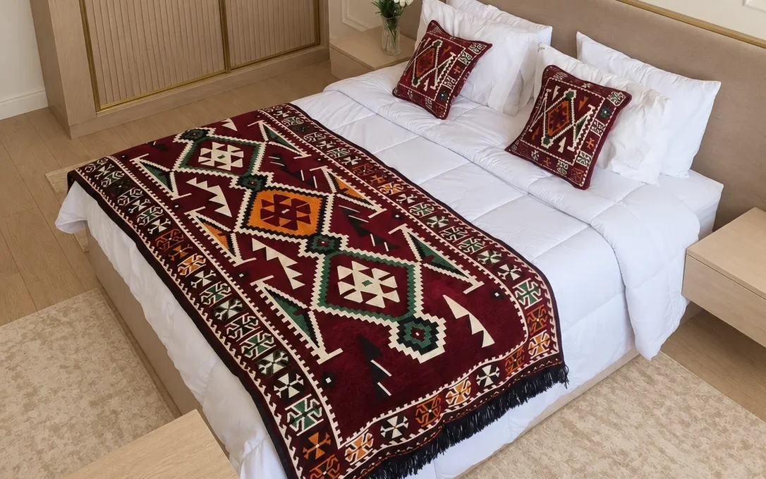 Burgundy kilim-pattern Moroccan bed runner rug with black fringe laid across the foot of a white bed, with two matching square cushions.