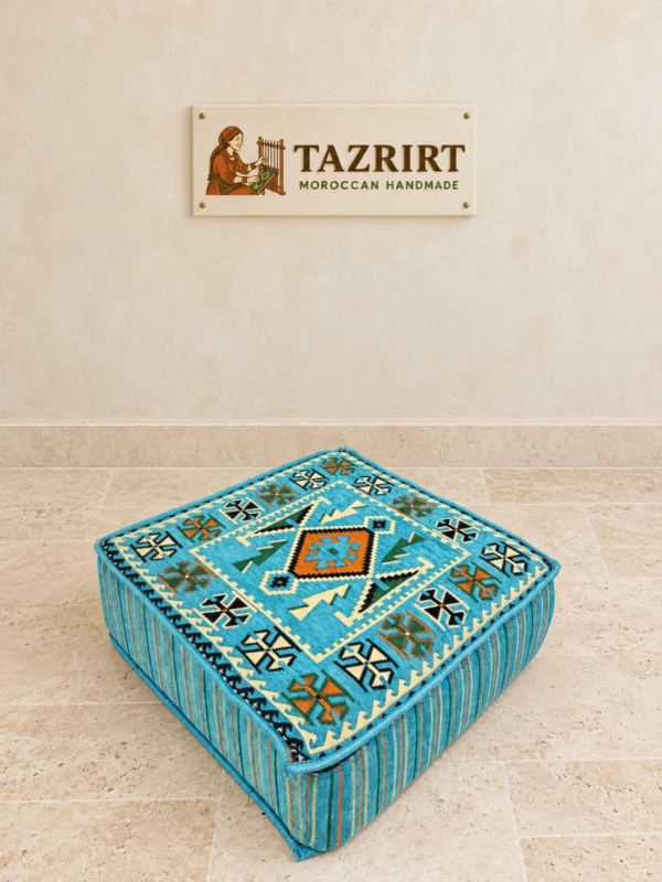 Turquoise kilim-pattern floor pouf with a rust-orange diamond at its center and striped, piped sides, on a stone floor.