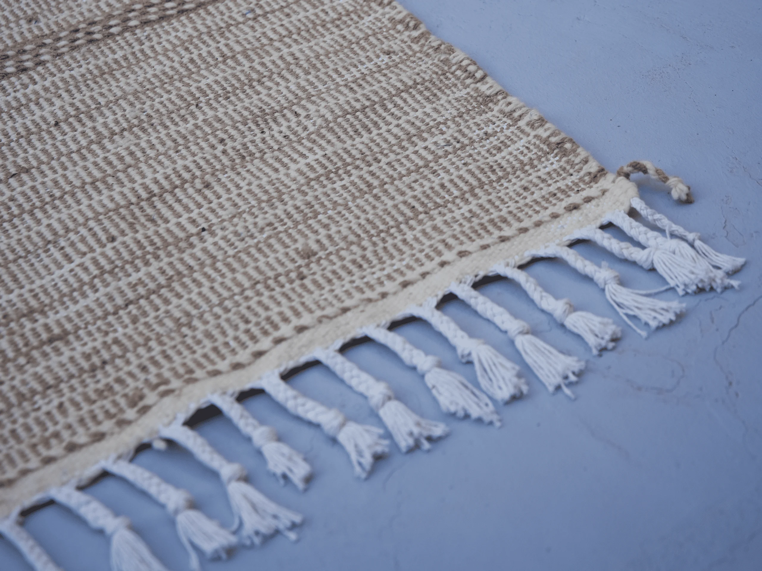 Close-up of a beige and ivory Zanafi wool runner showing woven texture, a finished border and braided ivory fringe.