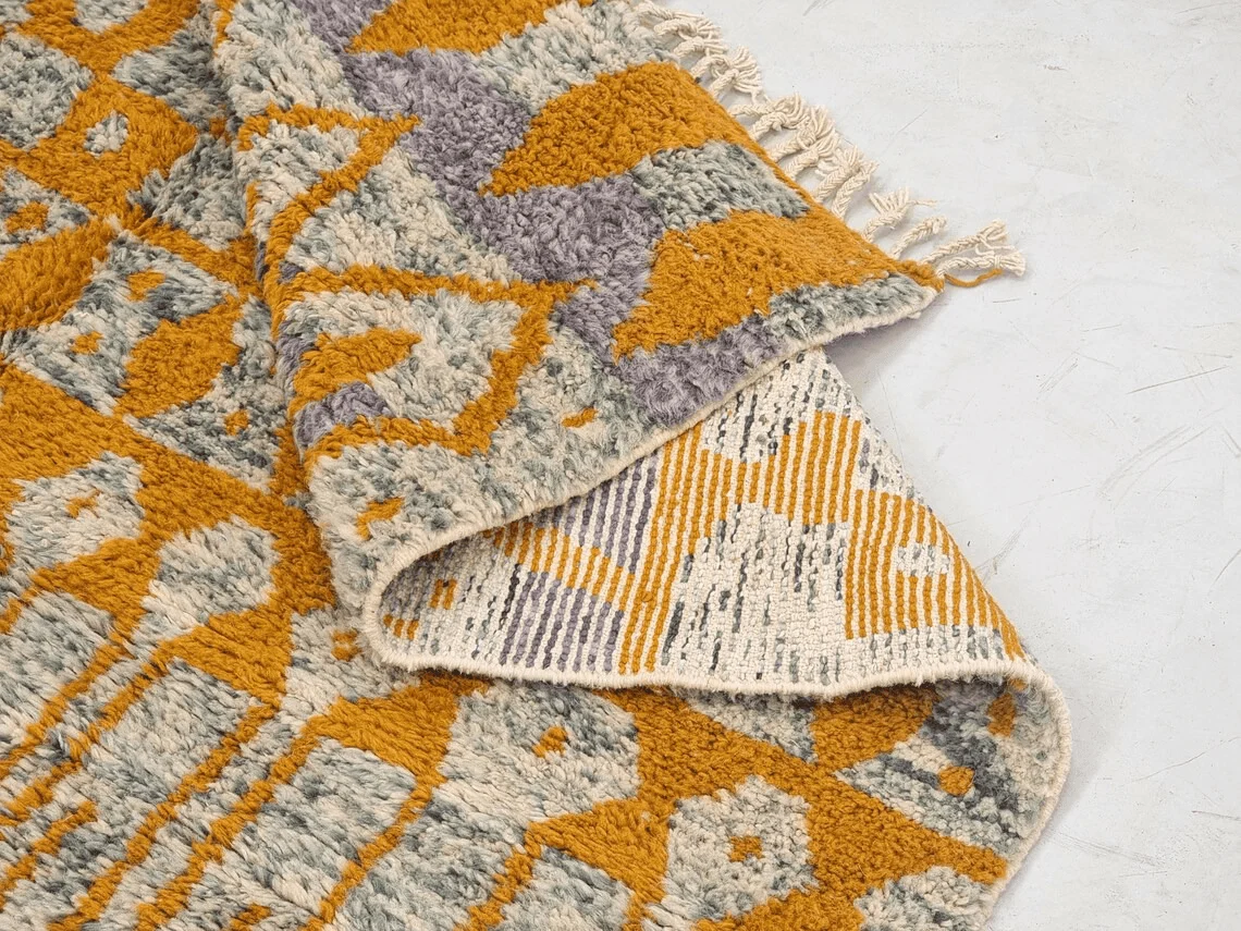 Folded corner of a mustard and gray Moroccan rug showing textured wool pile, the woven reverse and ivory fringe.