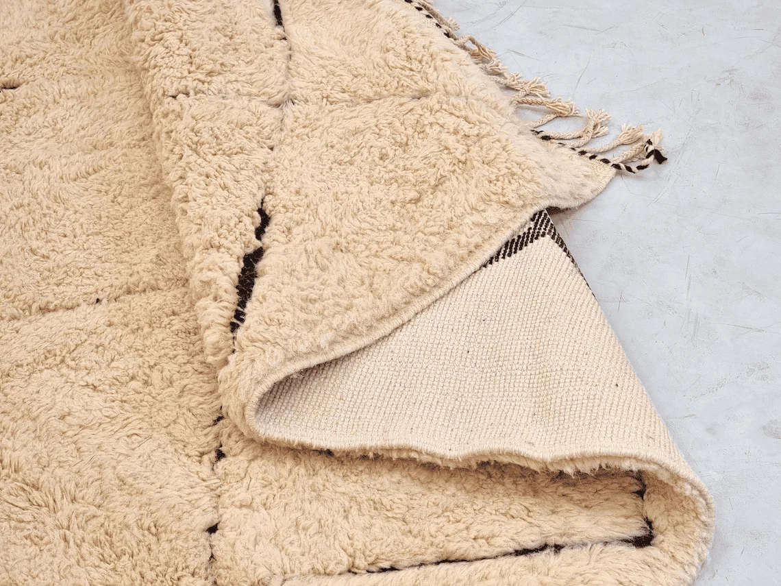 Close-up of thick cream wool pile and black diamond lines, with a folded corner revealing the woven underside and fringed edge.