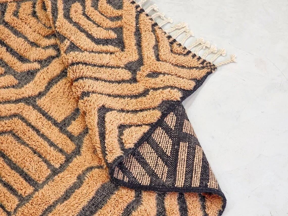 Close-up of raised peach wool pile between flat charcoal lines, with a folded corner and cream braided tassels.