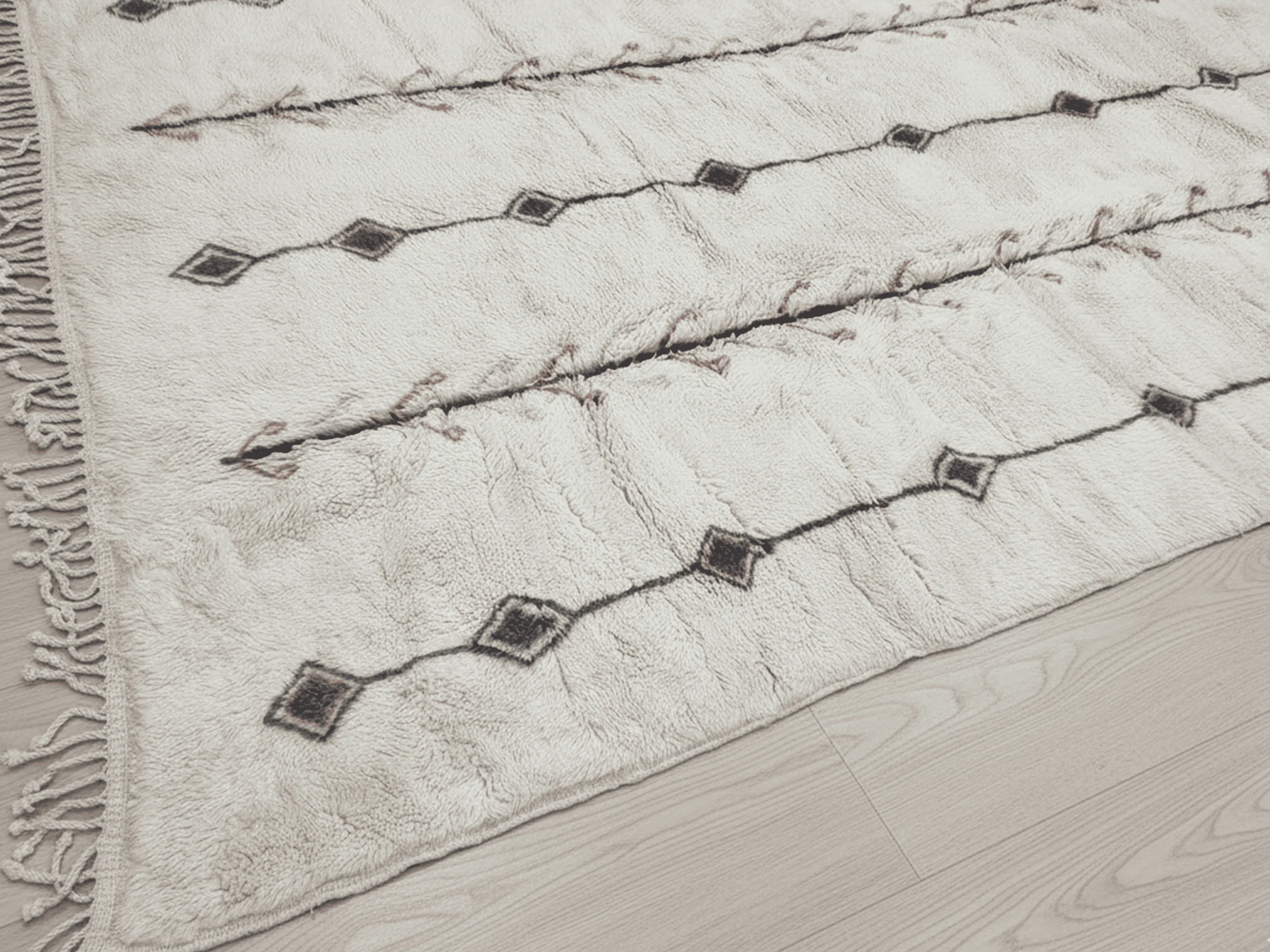 Close view of the rug's edge with charcoal diamond lines, rust branch marks and side fringe on a wood floor.