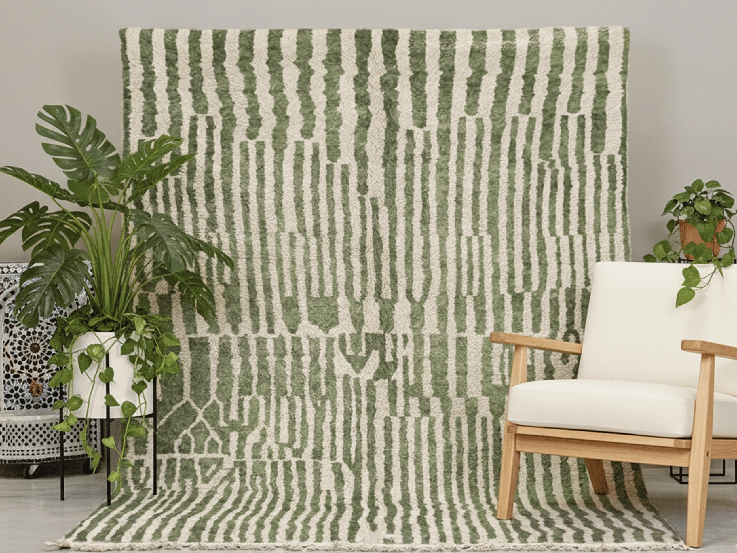 Green and ivory striped rug hung against a wall with a monstera plant and a cream armchair in front.