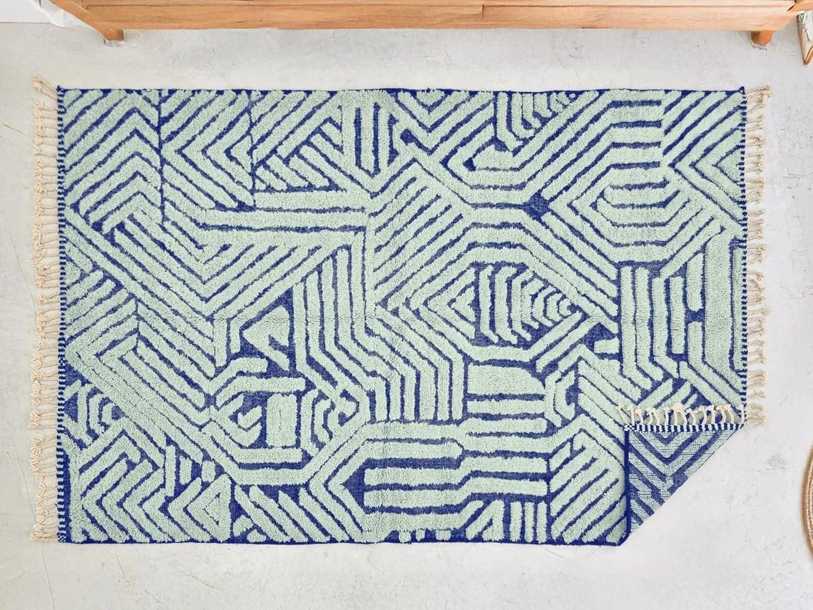 Top view of the mint and indigo geometric rug on a pale floor, with cream fringe and a folded corner showing the reverse.