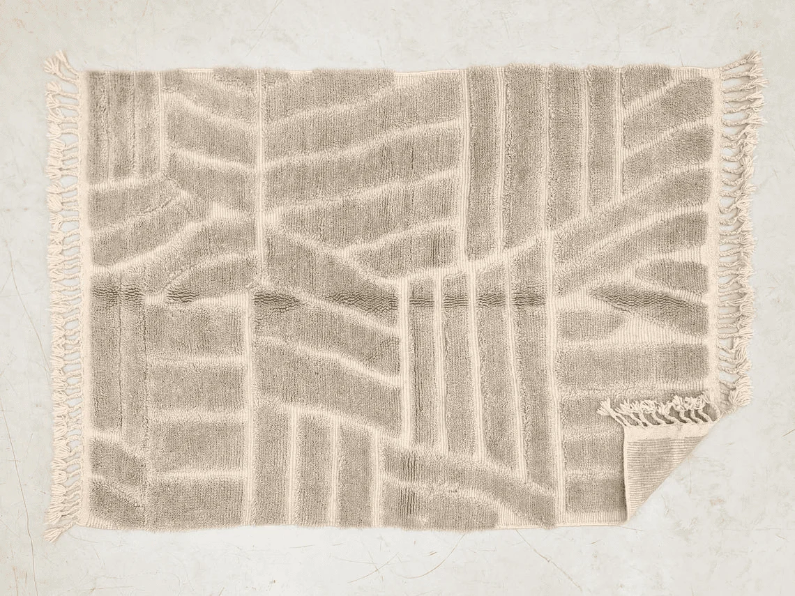 Top view of the gray and cream relief rug on a pale floor, with long fringe and one corner folded over.