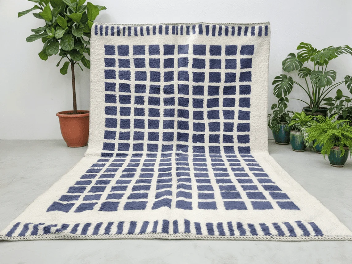 Ivory rug with indigo squares hung against a wall and spread on a concrete floor, with potted plants on both sides.