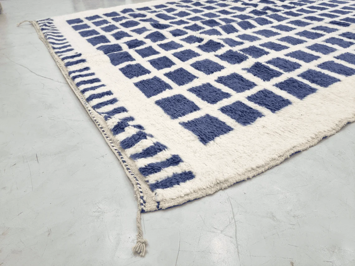 Corner of the ivory and indigo rug on a concrete floor, showing the dashed blue border and a braided tassel.