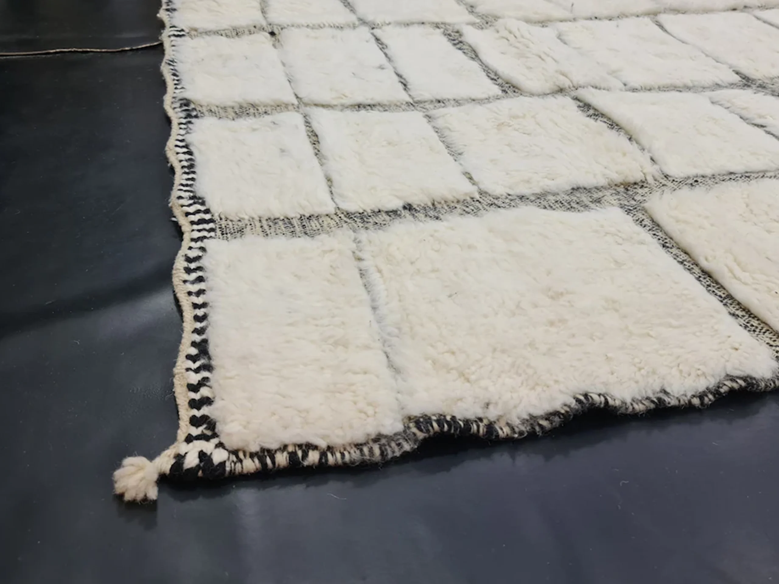 Corner of the ivory rug on a dark floor showing raised wool blocks, flat charcoal lines and a braided edge with a tassel.