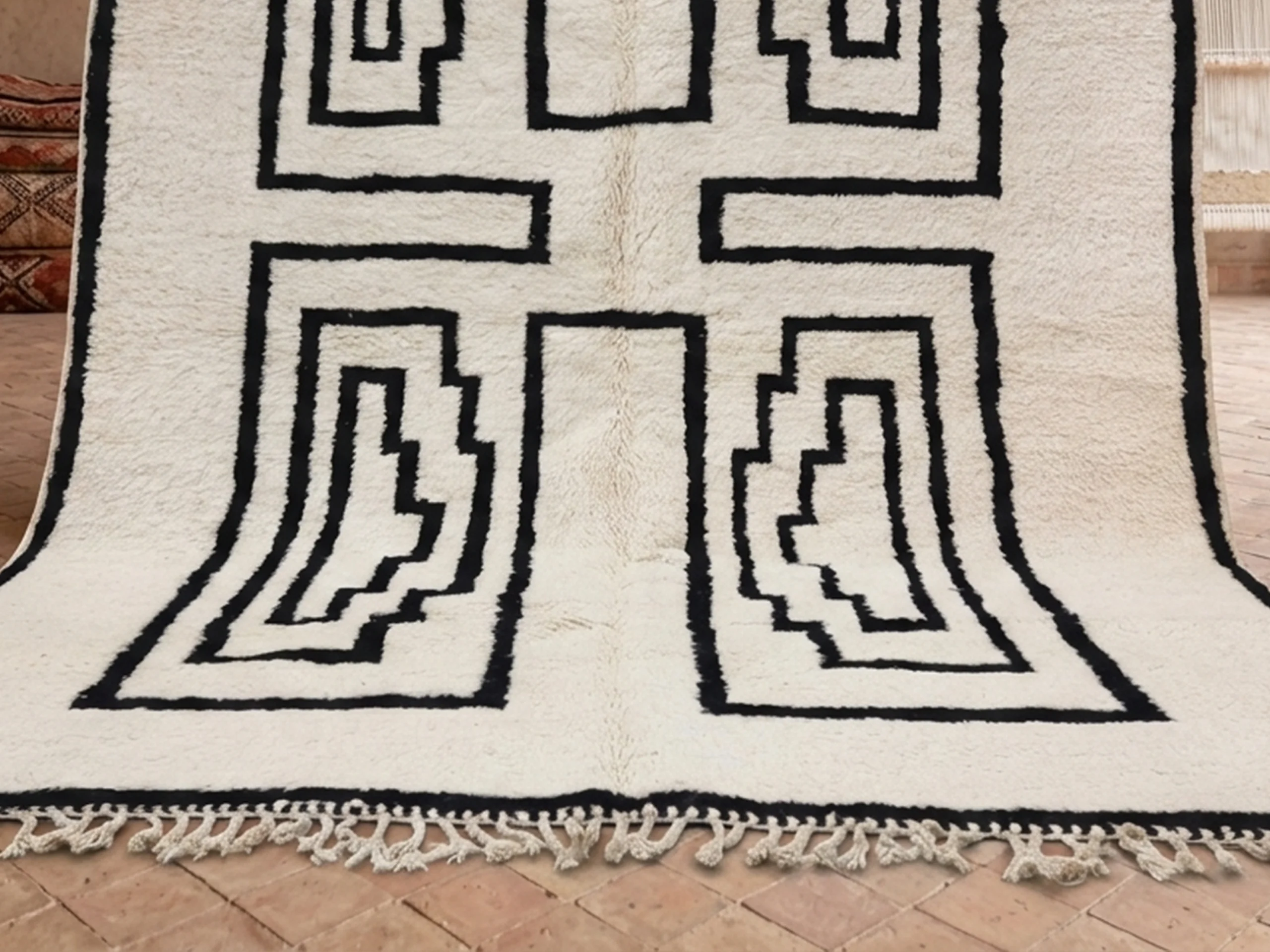 Lower half of the ivory and black rug hung on a wall, showing two stepped panels and fringe on a brick floor.