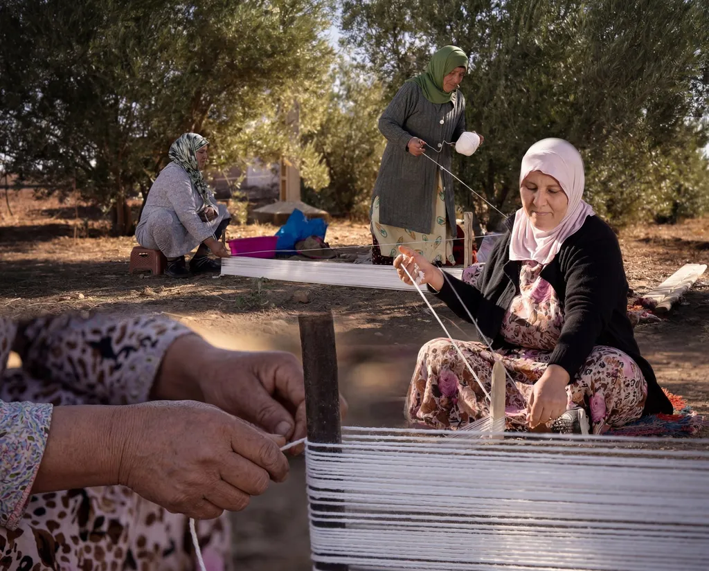 Women stretching white warp threads between wooden stakes on the ground.